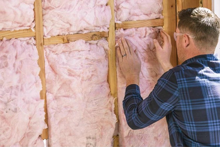 Man in flannel shirt installing fiberglass insulation in wall